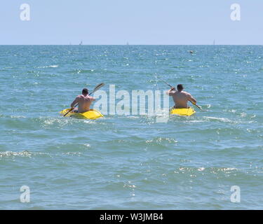 Deux hommes pagayant des kayaks jaunes, Boscombe, Bournemouth, Dorset, Angleterre, ROYAUME-UNI. 16th juillet 2019. Météo : magnifique jour d'été sur la côte sud avec une température dans les 20s basses, ce qui le rend très chaud sous le soleil chaud sur la plage. Deux hommes musclef pagayez furfurieusement des kayaks à la mer. Banque D'Images