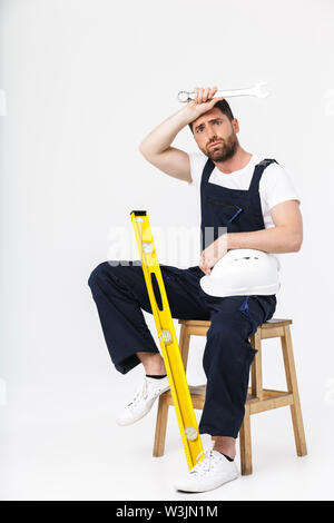 Toute la longueur d'un constructeur barbu fatigué homme portant une combinaison et casque assis sur une chaise isolée sur fond blanc Banque D'Images