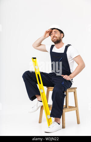 Toute la longueur d'un constructeur barbu fatigué homme portant une combinaison et casque assis sur une chaise isolée sur fond blanc Banque D'Images