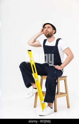 Toute la longueur d'un constructeur barbu fatigué homme portant une combinaison et casque assis sur une chaise isolée sur fond blanc Banque D'Images