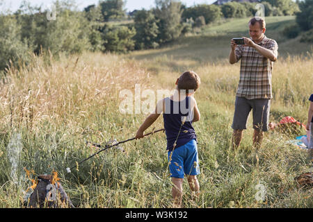 L'homme prend une photo de son fils le rôtissage de la guimauve sur un feu de camp. Famille de passer du temps ensemble sur un pré, près de la nature. Les gens sincères, du vrai momen Banque D'Images