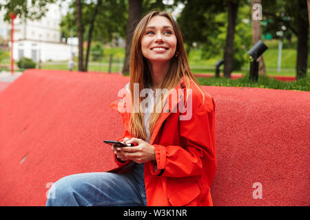 Cheerful attractive young girl wearing raincoat belle dépenses de temps à l'extérieur, à l'aide de mobile phone Banque D'Images