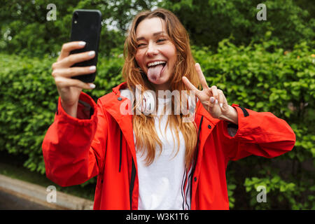 Image de jeune femme drôle 20s dans l'imperméable de couleur et souriant en tenant sur selfies portable tout en marchant à travers le parc vert sous la pluie Banque D'Images