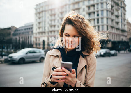 Laughing woman manteau beige à l'aide de smart phone standing in city près de road avec des voitures. La saisie des messages texte sur femelle appareil mobile à l'extérieur Banque D'Images