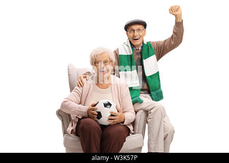 Cheerful senior homme et femme soccer fans avec un ballon de football et un foulard assise sur un fauteuil isolé sur fond blanc Banque D'Images