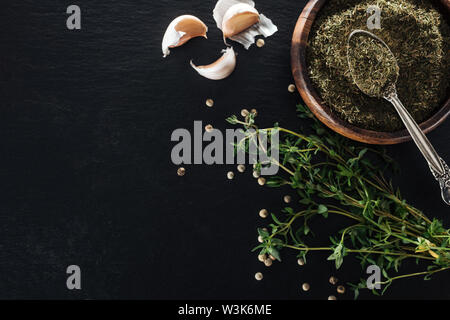 Vue de dessus de thym séché dans bol en bois avec cuillère en argent près de l'herbe verte, de poivre blanc et de gousses d'ail sur fond noir Banque D'Images