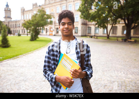 Un étudiant africain souriant avec des lunettes et des livres près de l'université. Portrait d'un jeune homme noir heureux debout sur un fond universitaire. Banque D'Images