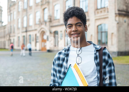 Un étudiant africain souriant avec des lunettes et des livres près de l'université. Portrait d'un jeune homme noir heureux debout sur un fond universitaire. Banque D'Images