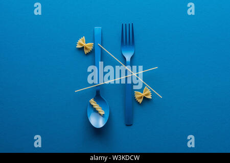Top View of spaghetti sur bleu cuillère en plastique à l'envers et la fourche près de deux sortes de pâtes sur fond bleu Banque D'Images
