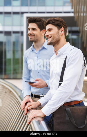 Photo d'un heureux jeune deux amis collègues hommes d'affaires à l'extérieur debout à côté holding phone. Banque D'Images