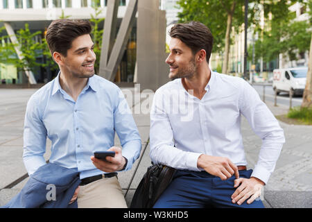 Image d'un jeune deux amis collègues hommes d'affaires assis près de centre d'affaires à parler les uns avec les autres à l'aide de téléphone mobile. Banque D'Images