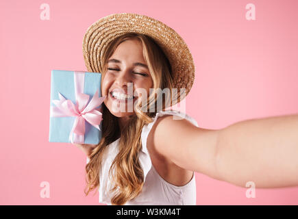 Photo de a smiling cute young Pretty woman posing isolé sur fond mur rose holding present gift box prendre une caméra par selfies. Banque D'Images