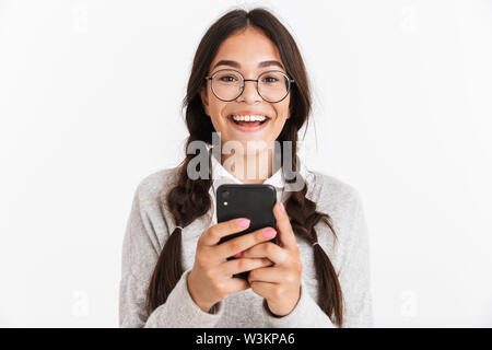 Gros plan Photo de surpris adolescente portant des lunettes et de l'uniforme scolaire smiling while holding smartphone isolé sur fond blanc Banque D'Images