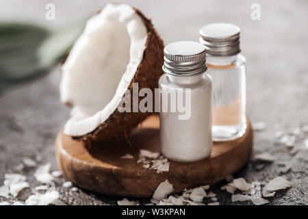 Focus sélectif des bouteilles avec des produits de beauté de coco sur planche de bois près de la moitié de la noix de coco sur fond texturé gris Banque D'Images