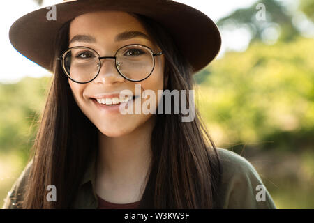 Photo de jeune femme joyeuse avec de longs cheveux noirs portant des lunettes et chapeau élégant balade dans Green Park, journée ensoleillée Banque D'Images