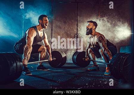 Deux barbus tattoed musculaire, l'entraînement des athlètes poids lourds de levage bar dans la fumée à la salle de sport. Scream. Travailler dur. Exercice pour les muscles du dos Banque D'Images