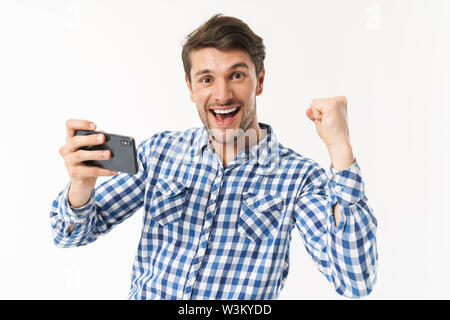 Photo de heureux l'homme à chemise décontractée jouer jeu vidéo sur cellulaire et un poing serrant isolé sur fond blanc Banque D'Images