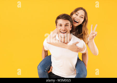 Portrait of content couple homme et femme t-shirts de base montrant signe de paix tout en faisant piggyback ride sur fond jaune isolé Banque D'Images