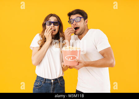 Portrait de couple choqué l'homme et la femme à lunettes 3D'être effrayé en se tenant debout avec seau de maîs éclaté sur fond jaune isolé Banque D'Images