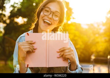 Photo d'une jeune fille enjouée d'étudiants portant des lunettes assis à l'extérieur dans la nature park holding livre. Banque D'Images