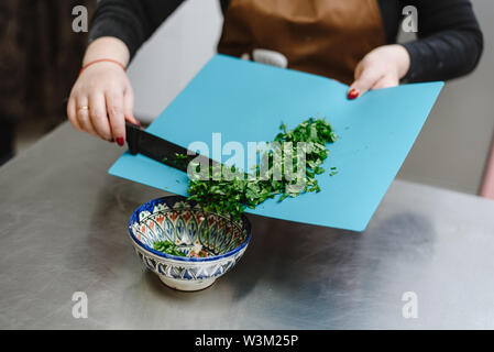 La jeune fille coupe verts, oignons, persil et divers assaisonnements avec un couteau sur une planche à découper. Femme cuire salade pour la cuisson. Banque D'Images