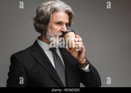 Image de l'homme d'adultes barbus portant costume noir officiel de boire du café à partir de la tasse de papier sur fond gris isolé Banque D'Images