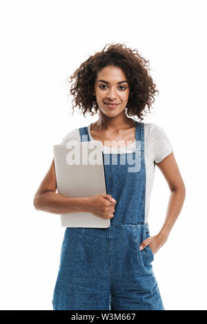 Image de young african american woman vêtus de salopettes en denim smiling, ordinateur portable isolé sur fond blanc Banque D'Images