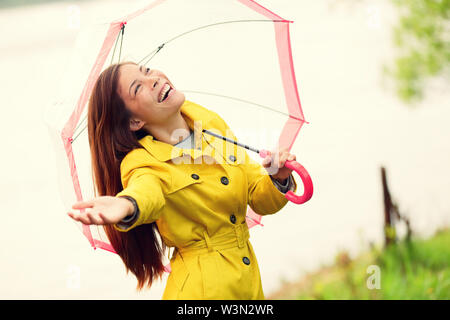 Automne femme heureuse après la pluie marcher avec un parapluie. Modèle féminin à la recherche jusqu'au ciel de compensation sur joyeuse journée d'automne pluvieuse portant imperméable jaune à l'extérieur dans la nature forêt par lac. Mixed Race girl. Banque D'Images