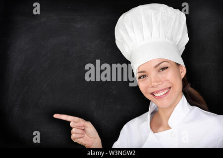 Pointage Chef showing blank blackboard menu. Happy female chef, cuisinier ou baker montrant l'affichage du menu tableau vide. Femme portant des uniformes blancs chef et un chapeau noir sur fond noir. Banque D'Images