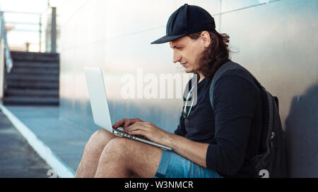 Un jeune homme aux cheveux longs et une casquette de baseball est assis sur une planche à roulettes jaune sur l'asphalte, s'appuyant le dos à un mur de granit gris et de travaux. Banque D'Images