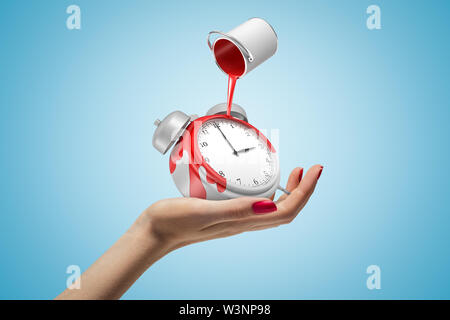 Femme hand holding réveil avec pot de peinture rouge à l'envers au-dessus sur fond bleu Banque D'Images