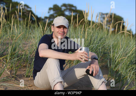 Heureux homme assis sur une dune de sable avec une tasse de café de voyage Banque D'Images