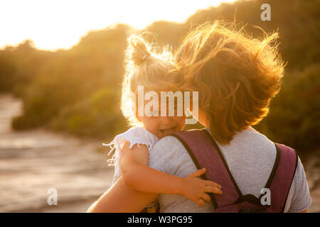 Portrait d'une jeune fille de race blanche de 3 ans aux cheveux blonds qui montre sa langue de manière ludique. Dans les bras de la mère, l'image en contre-jour au coucher du soleil. Banque D'Images