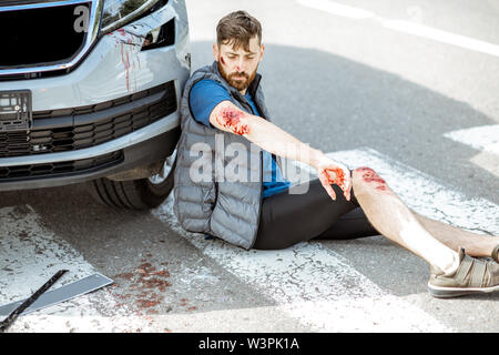 Homme blessé avec des plaies saignantes assis sur le passage pour piétons près de la voiture après l'accident de la route Banque D'Images