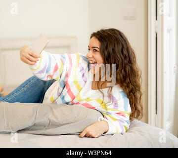 La mode jeune adolescent excité de prendre une femme sur selfies smart mobile phone sur le lit chez lui. Heureux de parler et de poster en ligne dans les médias sociaux apps. Dans le ne Banque D'Images