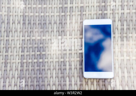 Reflet de ciel bleu avec des nuages dans le téléphone-smartphone, qui se trouve sur une table en osier clair contre l'arrière-plan. Le flou et l'abstraction Banque D'Images