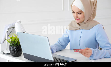 Concept d'achat en ligne. Portrait de jeune femme musulmane en beige hijab et traditionnelle robe bleue est l'achat en ligne avec une carte de crédit et l'ordinateur portable. Banque D'Images