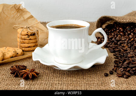 Vue avant du café noir en blanc chaud tasse soucoupe, élégant avec des biscuits sucrés et de dispersion des grains de café sur fond d'un sac en milieu rural. Banque D'Images