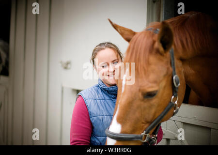 Jeune femme debout à côté de son cheval brun qui dépasse sa tête par la porte ouverte de la stabilité. Banque D'Images