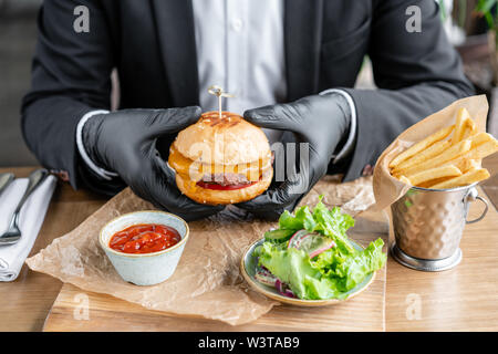 Jeune homme en costume et gants noirs manger de délicieux juteuse cheeseburger avec beef chop. Des hamburgers dans le café avec salade et frites, restauration rapide. Banque D'Images