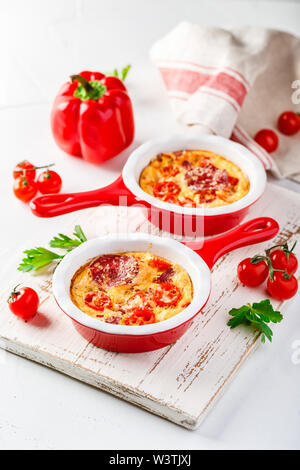 Frittata aux poivrons doux frais, tomates et de salami. Omelette italienne en céramique rouge poêlons sur un fond blanc. Close up Banque D'Images