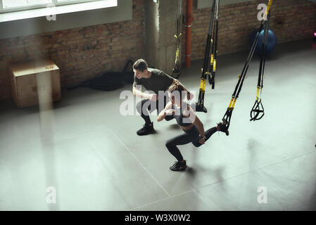 L'accroupissement profond. Jeune couple actif dans les jambes avec la formation de sport fitness Trx attaches, au sport. Entraîneur personnel. Formation TRX. Ensemble de l'exercice Banque D'Images