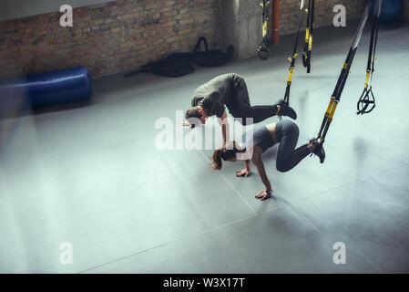 Entraînement intensif. L'homme et de la femme dans les vêtements de sport faisant de l'exercice dans la salle de sport TRX. Les jambes de formation de remise en forme avec salle de fitness à sangles Trx. Entraîneur personnel. Formation TRX. Ensemble de l'exercice Banque D'Images