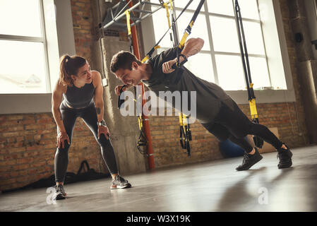 Donner la motivation. Les jeunes et l'homme fort dans les vêtements de sport faisant pousser ups avec trx sangles et de remise en forme tout en souriant son entraîneur personnel est debout près de lui. Formation TRX. L'exercice ensemble. Couple actif Banque D'Images