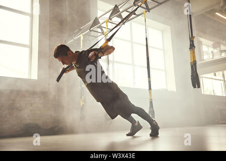 Push-up parfait. Vue latérale du jeune homme athlétique et solide dans les vêtements de sport faisant pousser ups avec trx sangles remise en forme à la salle de sport. La remise en forme. Formation TRX. Concept d'entraînement Banque D'Images