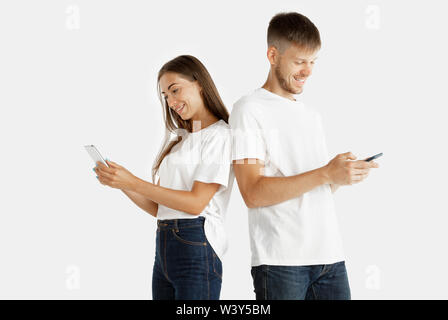 Beau jeune couple studio portrait isolé sur fond blanc. L'expression du visage, des émotions humaines, concept de publicité. L'homme et de la femme à l'aide de smartphones pour chating, Paris, regardant la vidéo. Banque D'Images