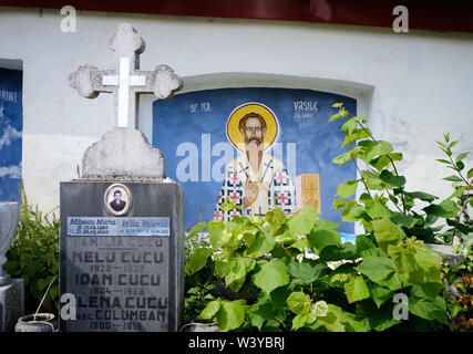 Façade mur peint avec image d'un saint dans un cimetière orthodoxe roumaine, encadrée par des croisements de tombstone et buissons croissant Banque D'Images