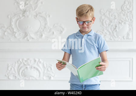 Un petit garçon avec des lunettes et un t-shirt bleu est en train de lire un livre dans une couverture verte avec intérêt. Banque D'Images