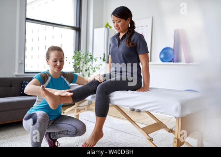 Caucasian woman physiothérapeute strectching la jambe et du genou d'un mid-adult chinese female patient assis sur une table de massage Banque D'Images