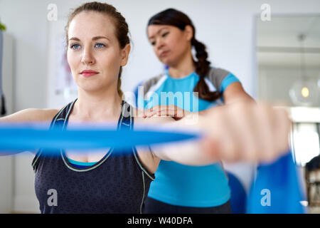 Femme chinoise professionnel physiothérapie donnant un traitement par l'utilisation de bandes de résistance élastique pour une jolie blonde client dans un bureau médical lumineux Banque D'Images